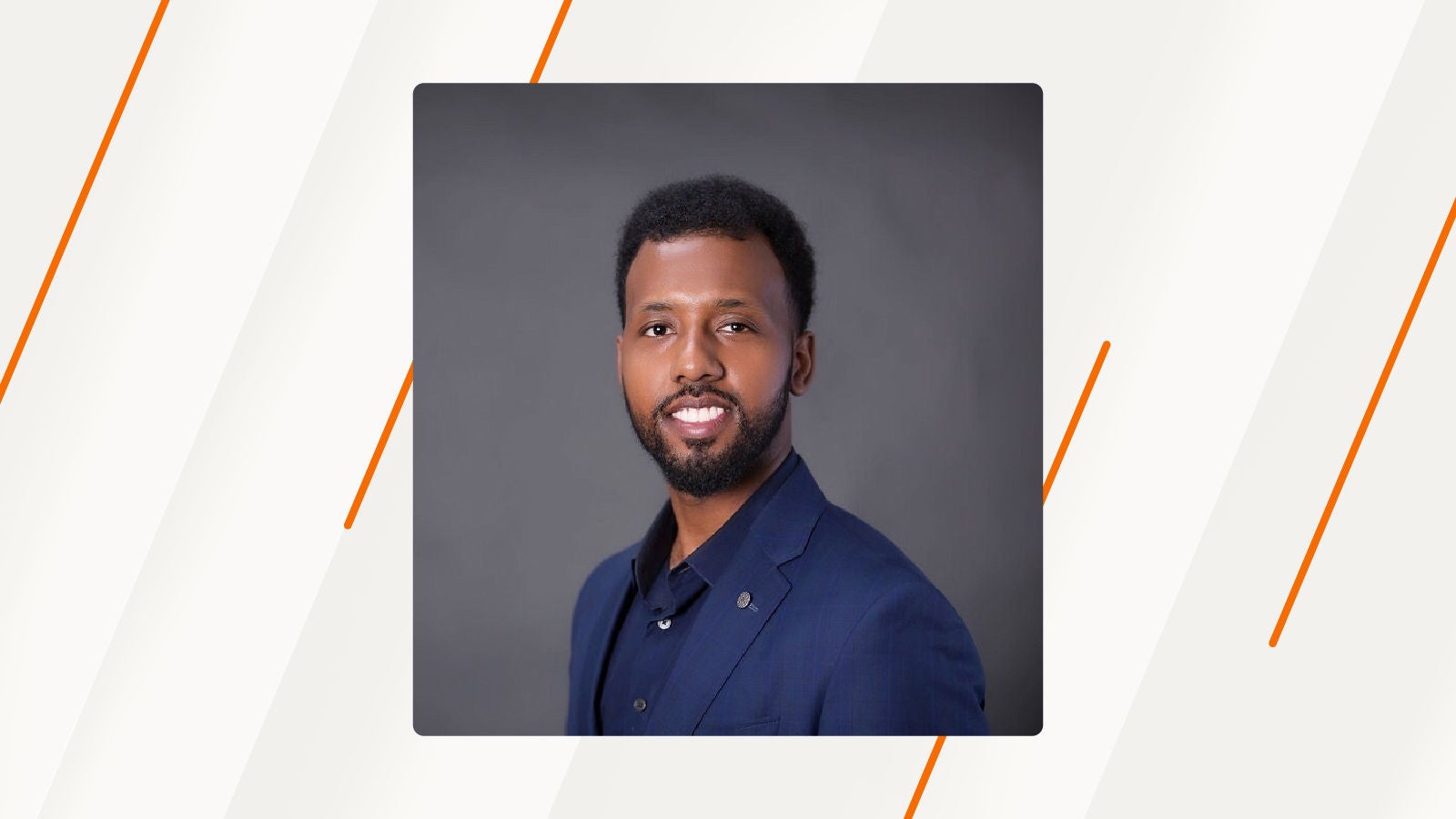 Portrait of a smiling man in a dark blue suit against a gray background, shown on a white and orange patterned backdrop.