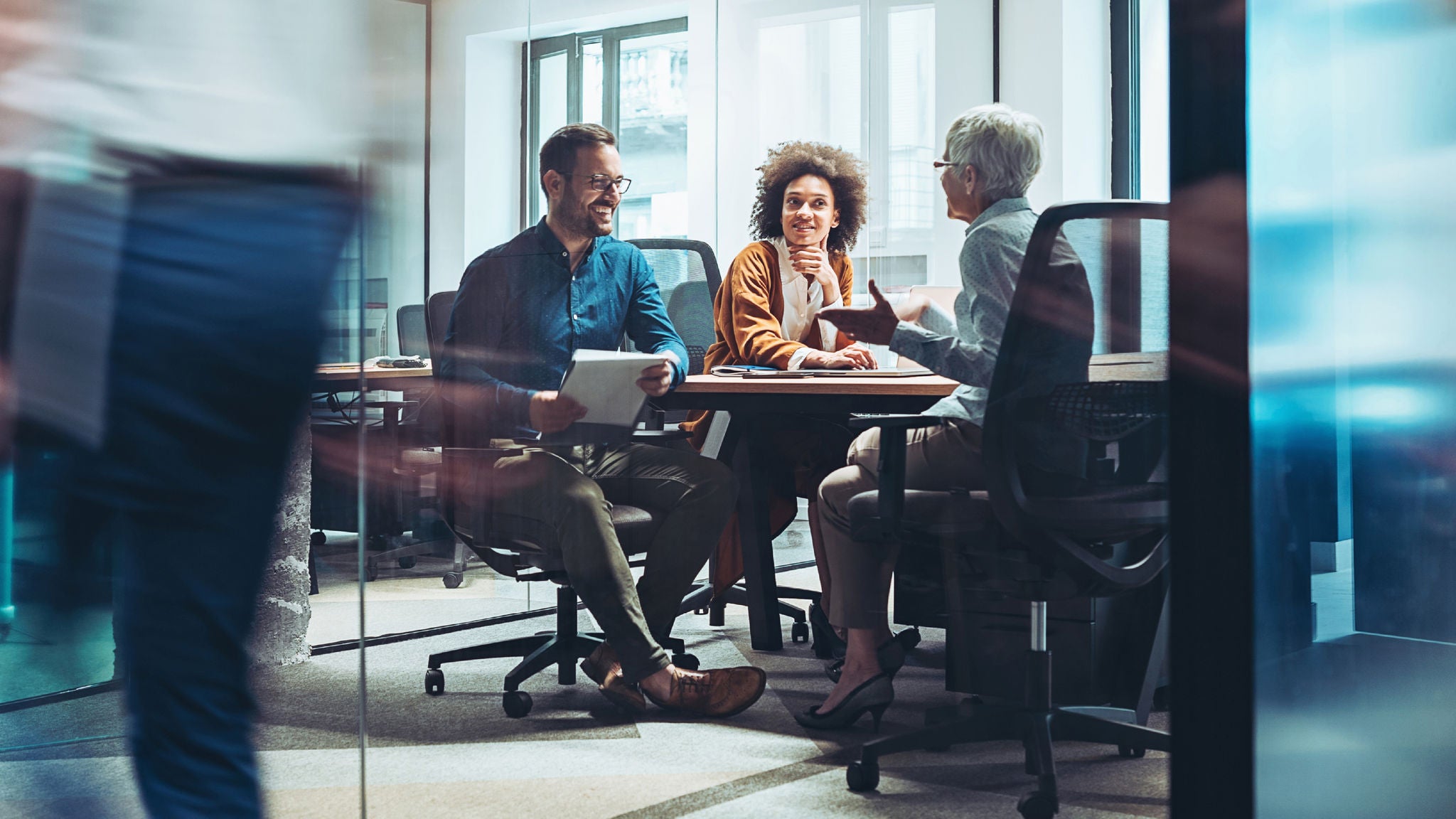 Three professionals seated around a meeting table in a modern office, engaged in a lively discussion with documents in hand, viewed through glass walls.
