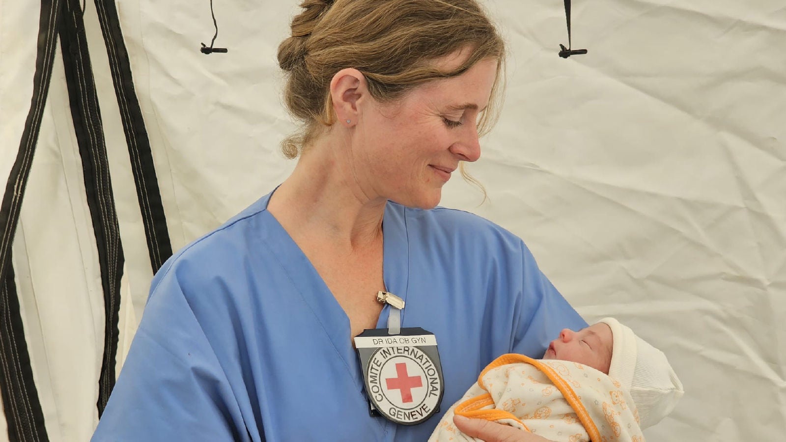 An ICRC medical worker in blue scrubs smiles while holding a newborn baby wrapped in a blanket inside a tent.