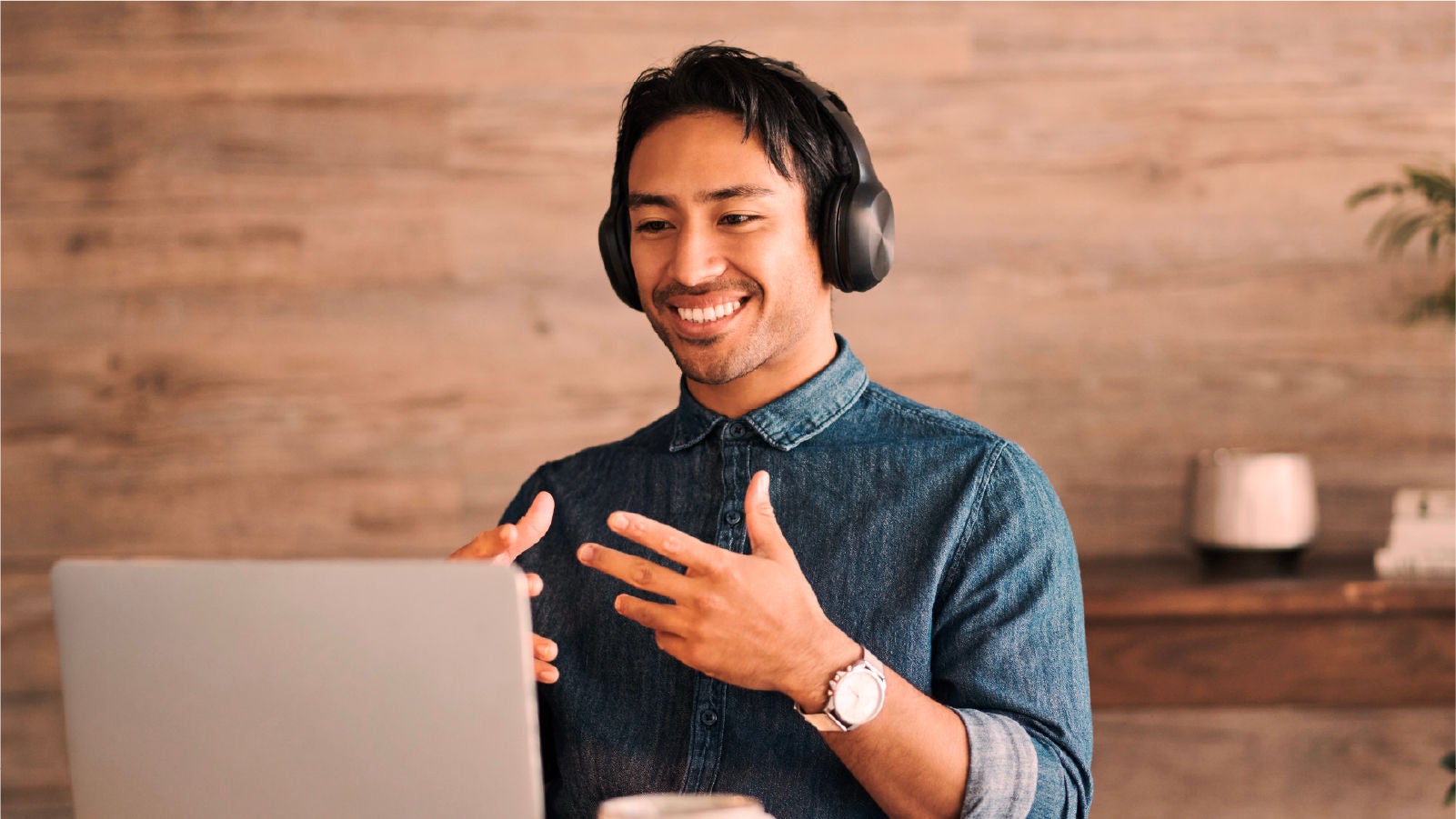 A man wearing headphones speaking and gesturing while using a laptop during a virtual meeting.