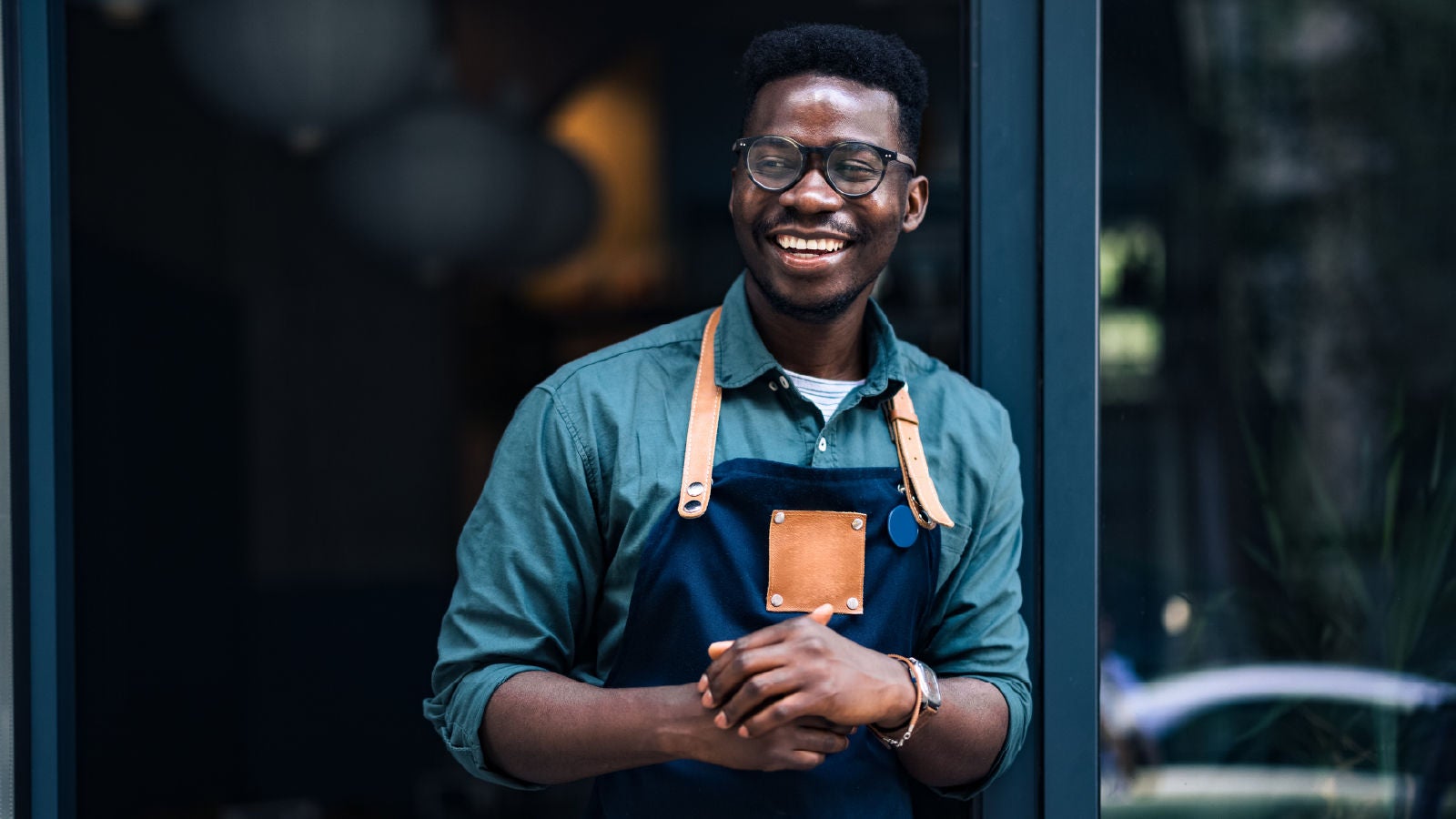 A smiling man wearing glasses and a green shirt with a navy apron stands outside a shop entrance.