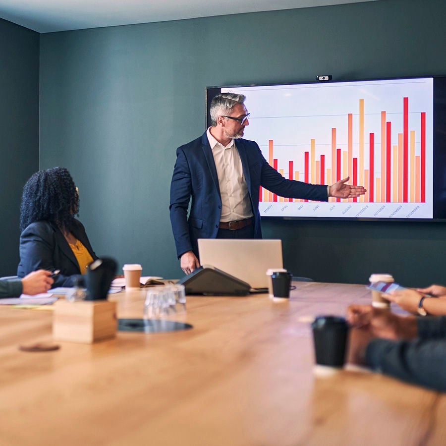 A business meeting where a presenter points to a bar chart on a wall-mounted screen while colleagues sit around a conference table with laptops and coffee cups.