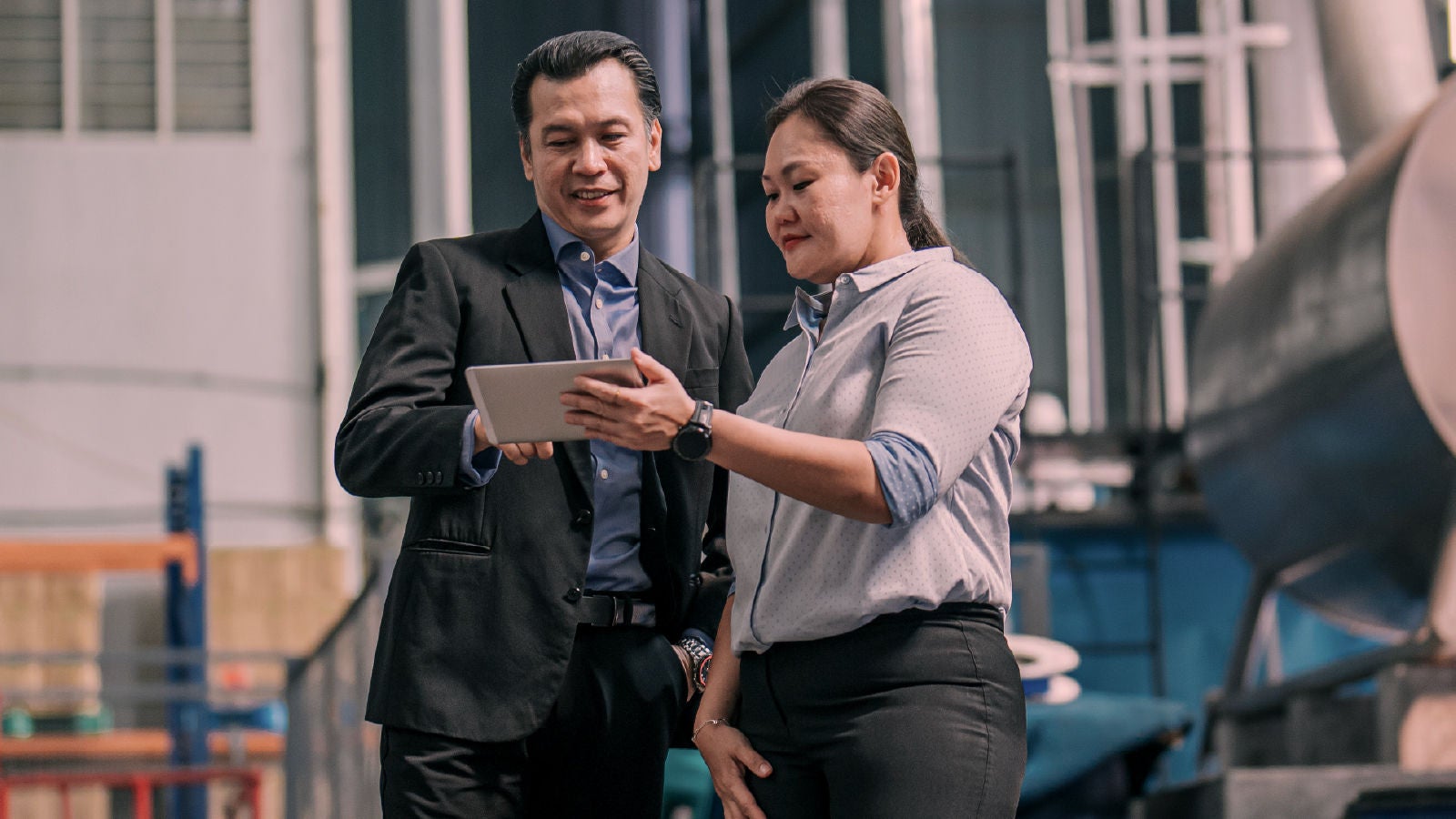 Two professionals standing in an industrial warehouse reviewing a tablet together.