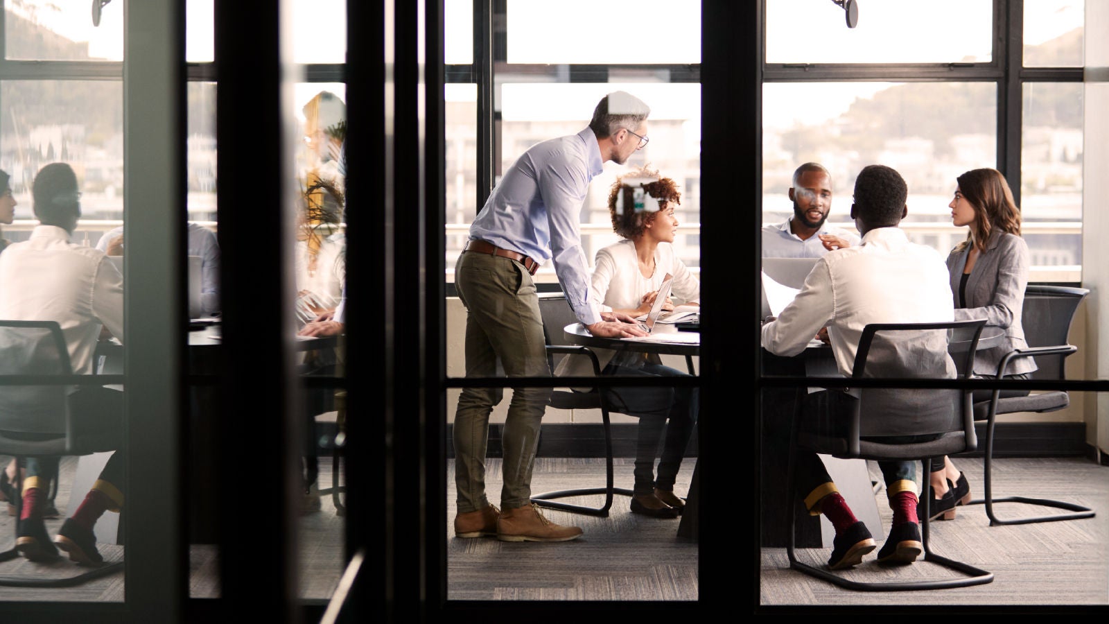 Five professionals in a glass-walled meeting room engaged in discussion around a table, with one person standing and leaning in to contribute.