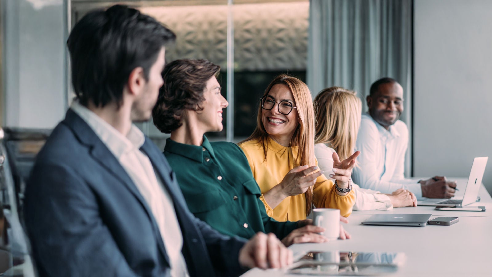 A group of professionals seated at a long table, talking and smiling during a meeting, with laptops and coffee cups placed in front of them.