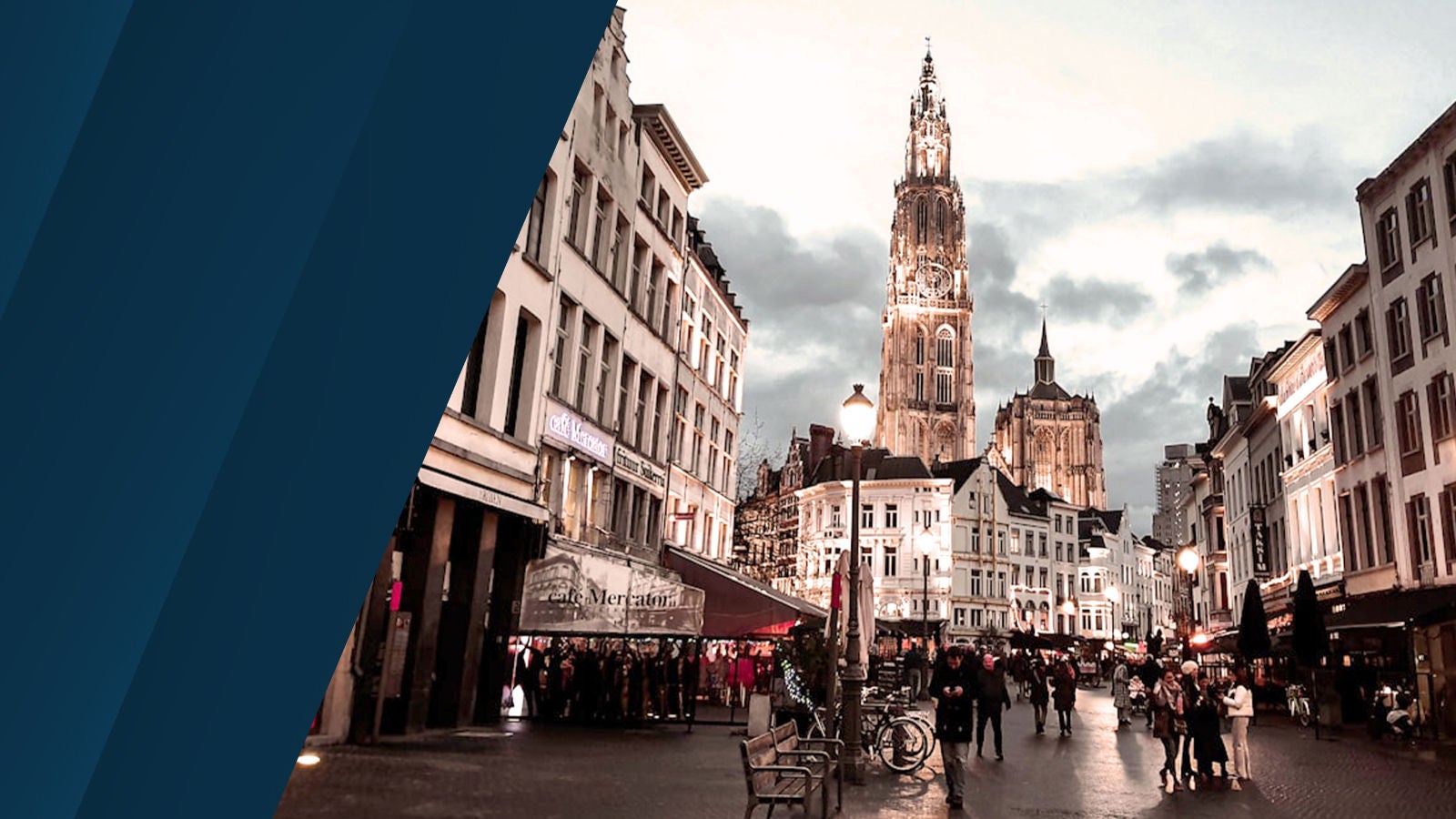 A bustling Belgian city street at dusk with people walking near shops and restaurants, framed by a towering illuminated cathedral under a moody sky.