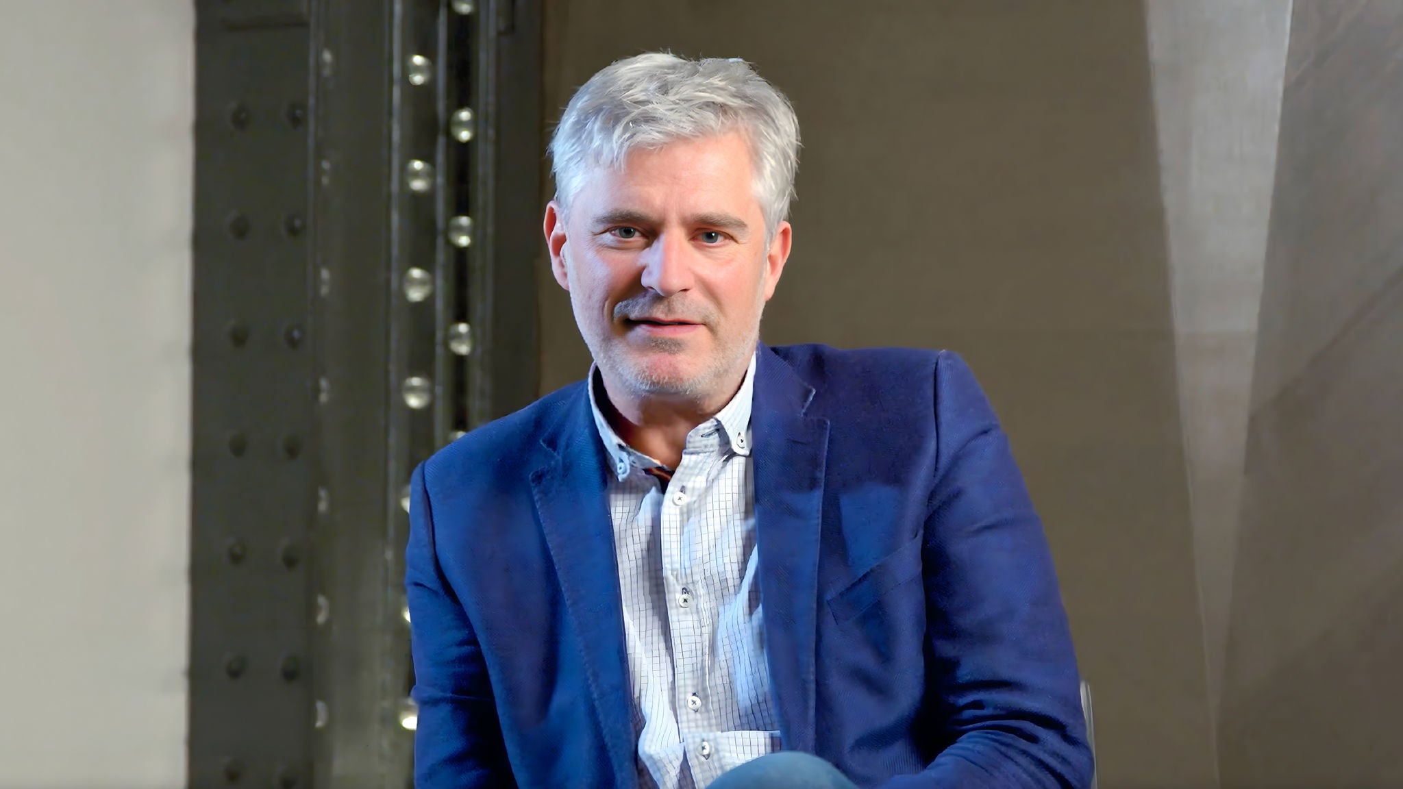 Man in a blue blazer sitting indoors during an interview or presentation.
