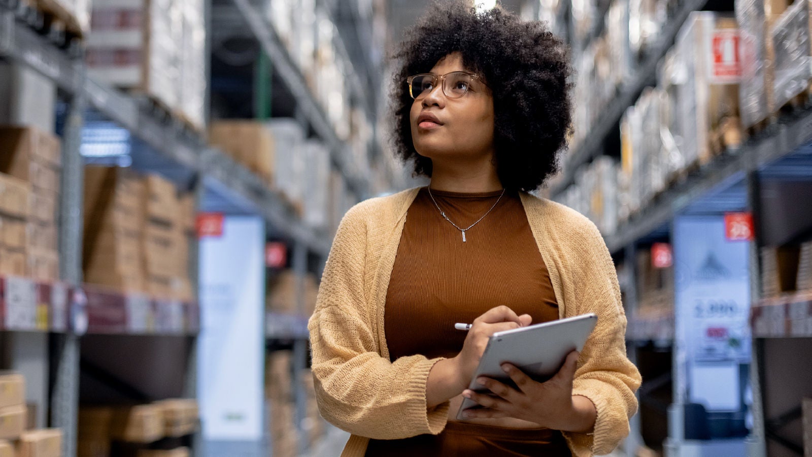 A woman with curly hair and glasses holds a tablet while standing in a warehouse aisle, surrounded by tall shelves of boxes.