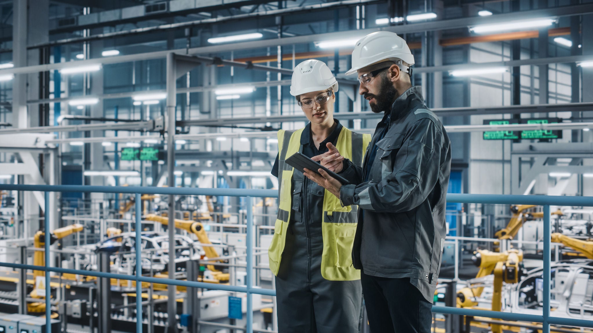Two engineers wearing hard hats and safety vests examining data on a tablet inside a modern factory with machinery in the background.