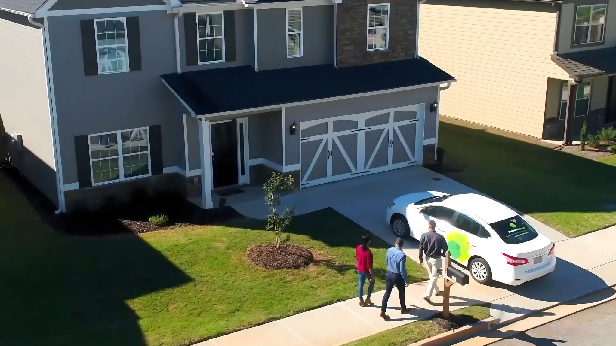 Aerial view of a suburban home with three people walking toward the front door from a parked car.