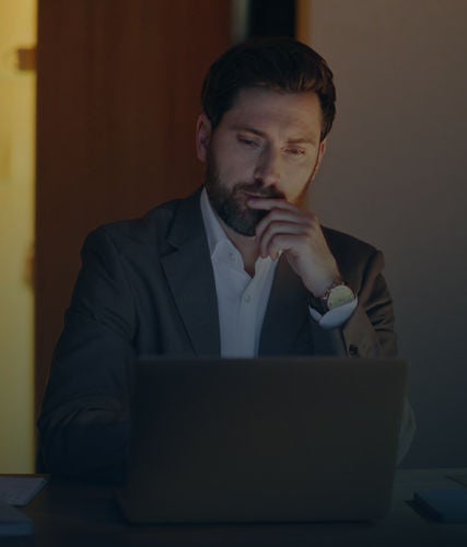 Business professional reviewing a laptop at a desk in a dimly lit office.