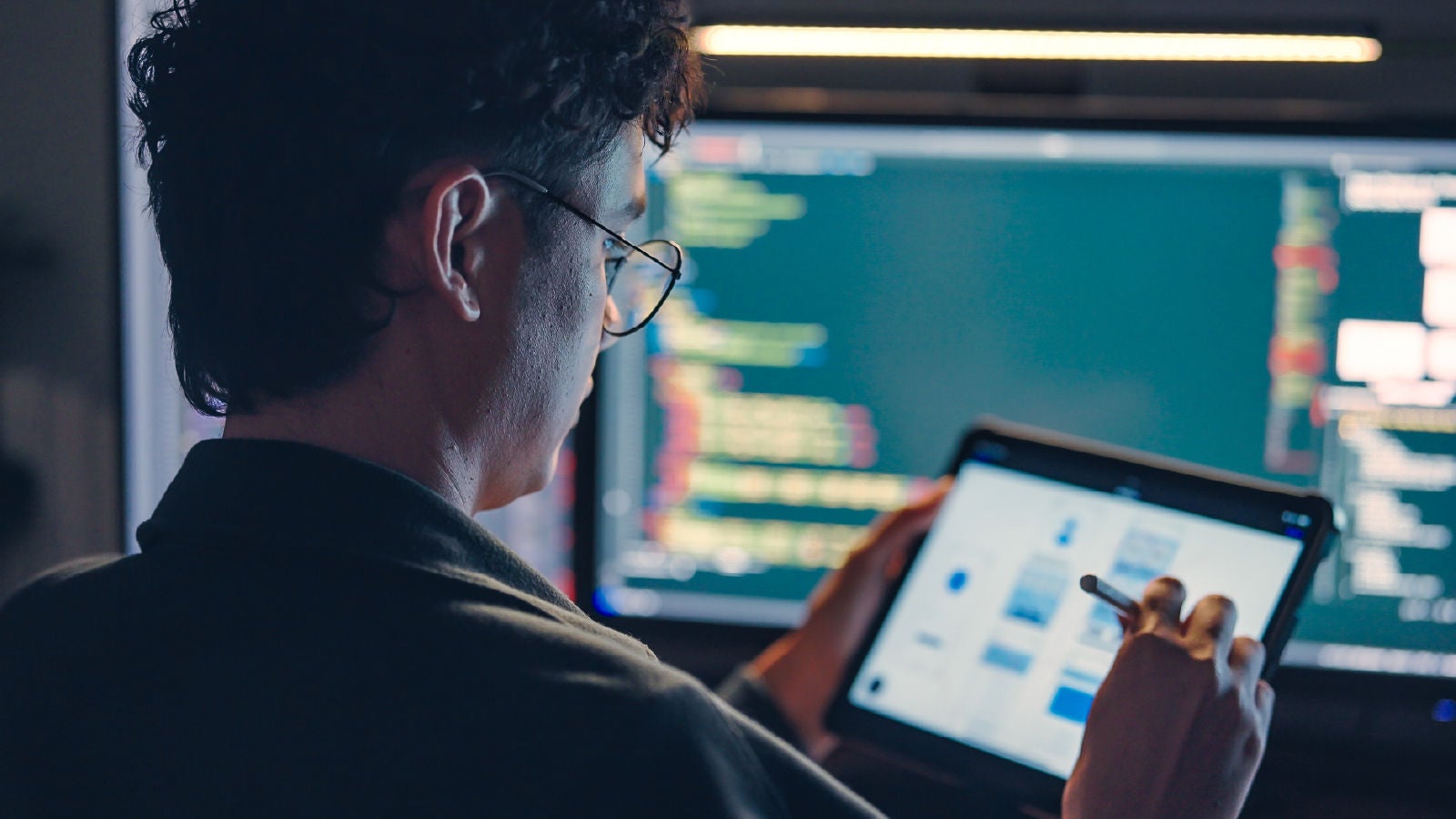 A professional reviewing data on a tablet while seated at a workstation, with multiple computer monitors displaying code and analytics in the background.