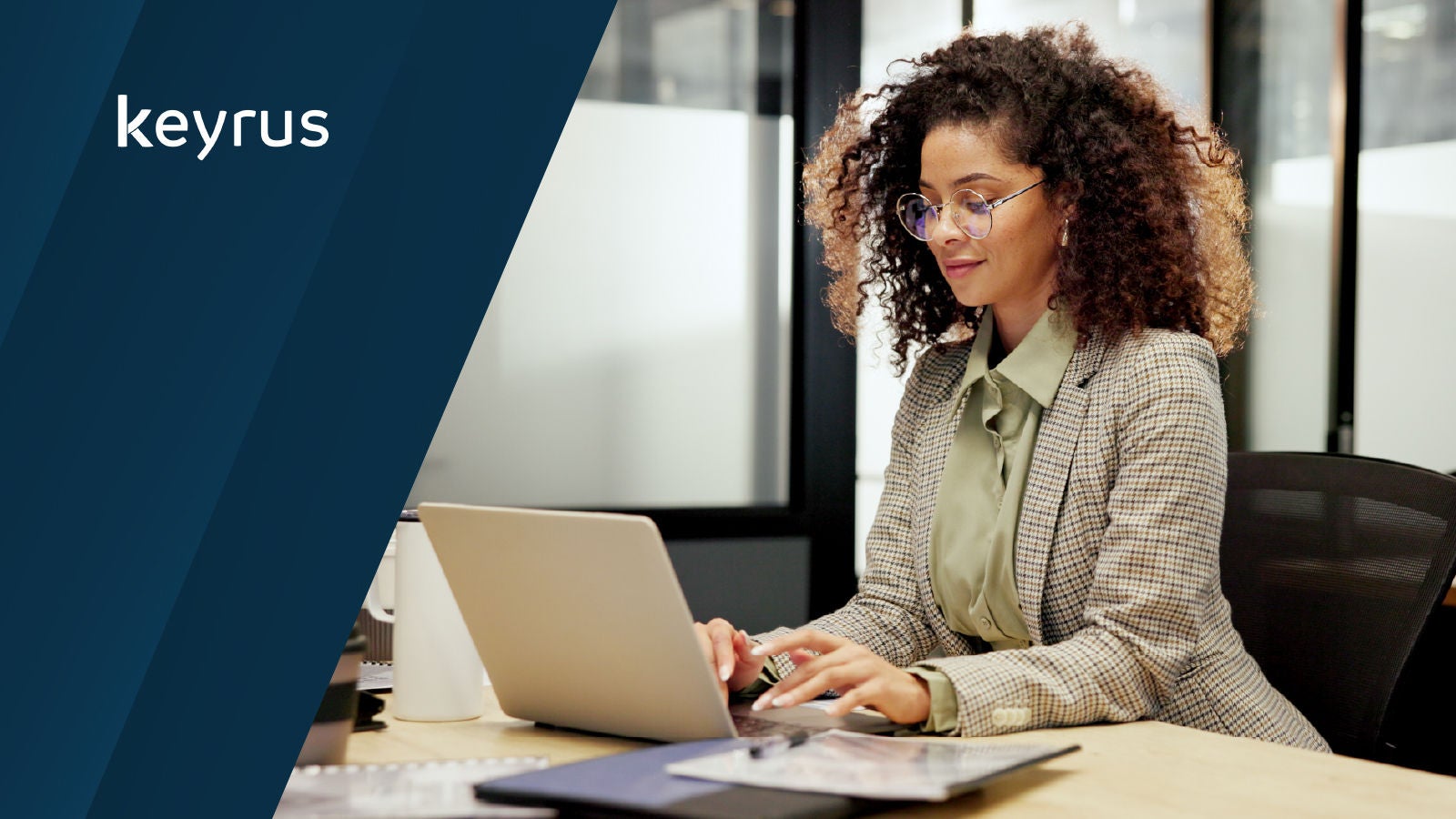 Keyrus logo displayed in the upper left. A woman with curly hair types on a laptop at a desk in a modern office environment.