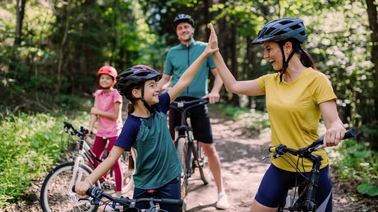 Family wearing bicycle helmets pauses on a wooded trail as a mother and child exchange a high five.