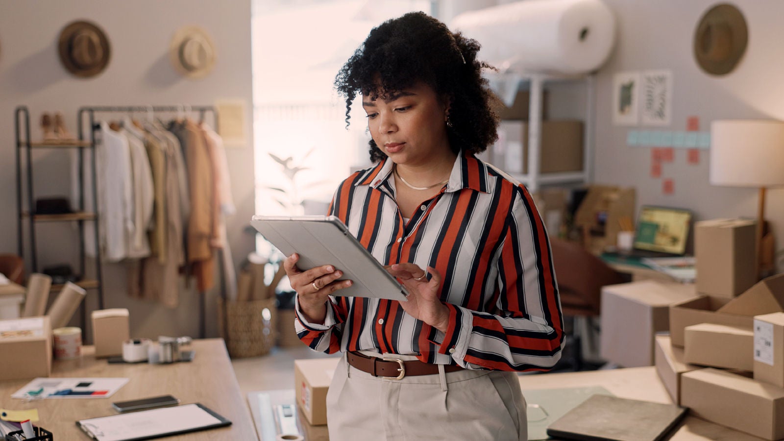 A woman in a striped shirt holding a tablet and reviewing information in a retail workspace with clothing racks and packaging materials in the background.