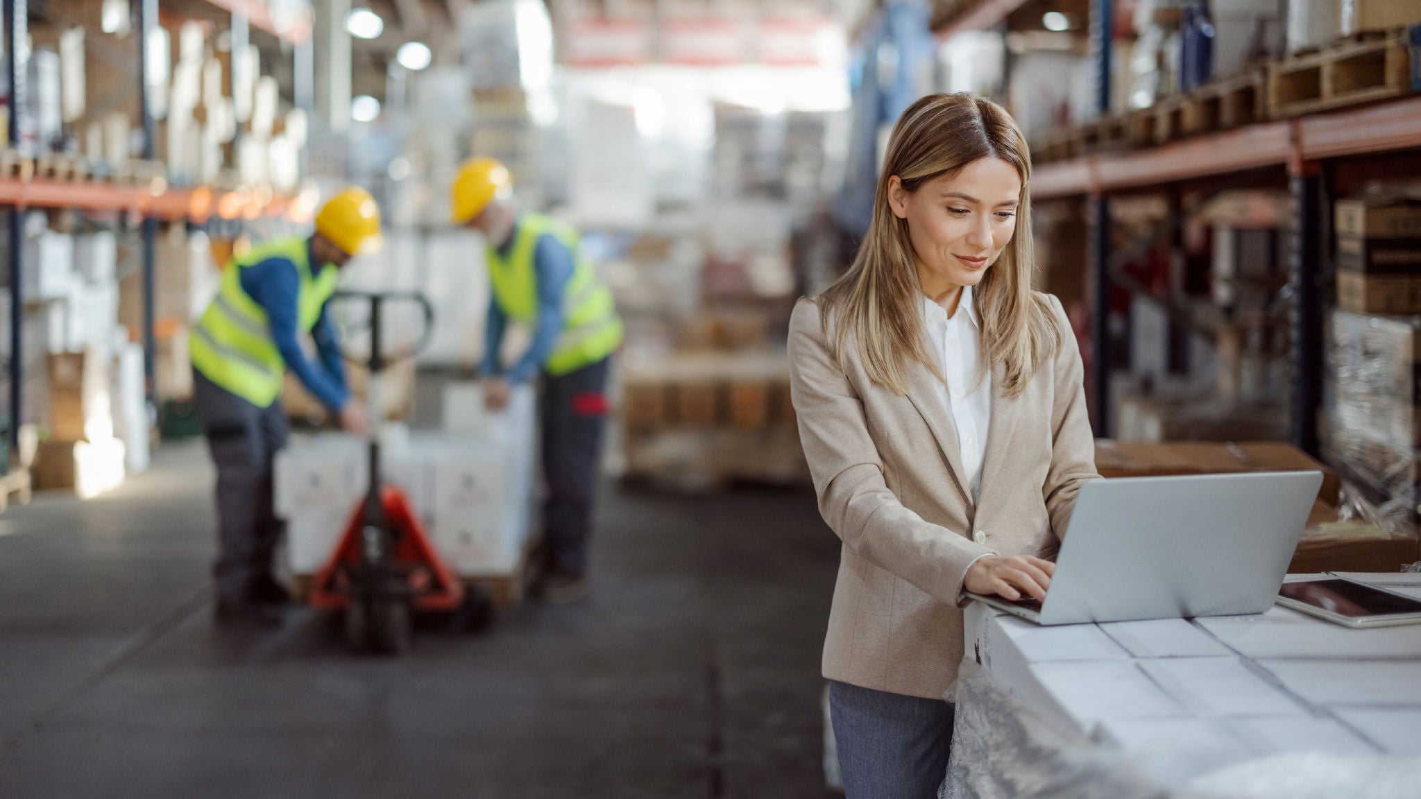 View of the warehouse with the woman working on her laptop in the foreground, while two workers in high-visibility vests are moving packages among tall shelves filled with inventory.