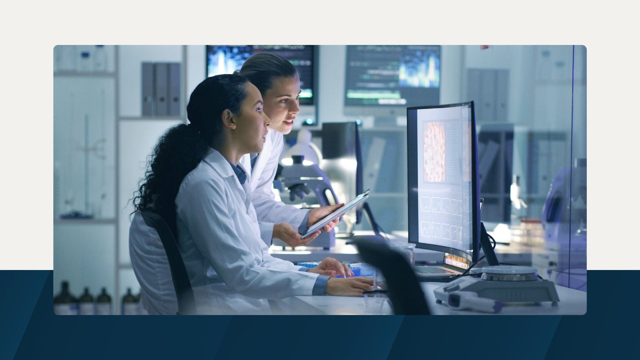 Two scientists in lab coats working together at a computer station in a modern laboratory, surrounded by monitors displaying scientific data.