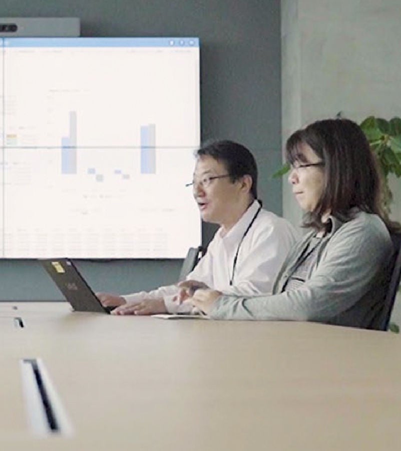 Two business professionals collaborating in a meeting room with a large digital screen displaying analytics.
