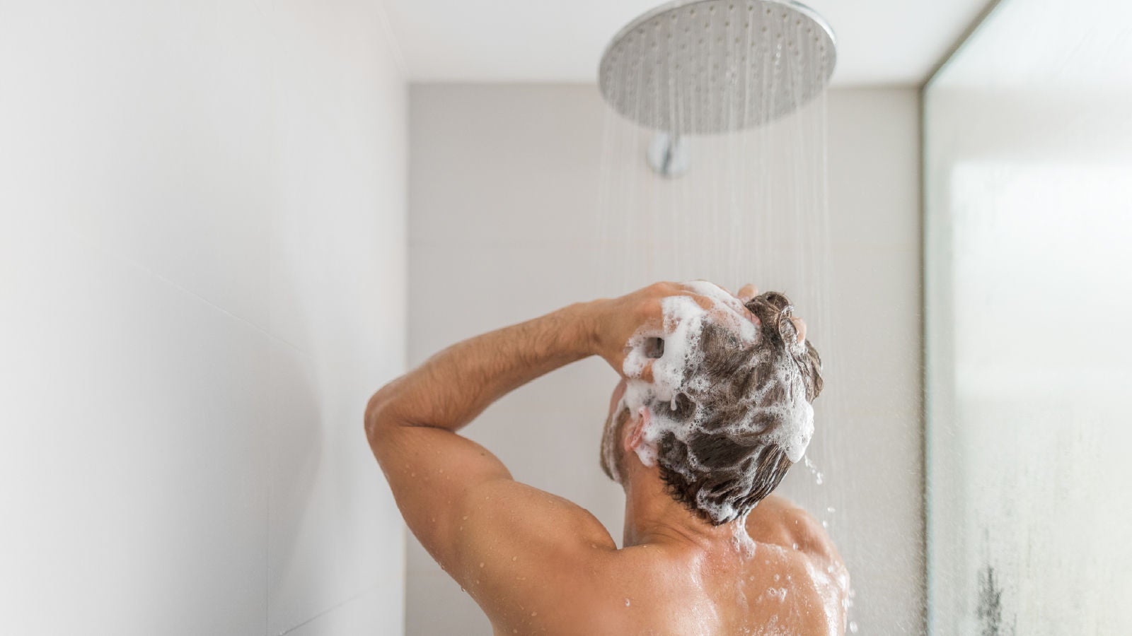 A man washing his hair under a showerhead, with soap lather and running water, representing cleanliness and freshness.