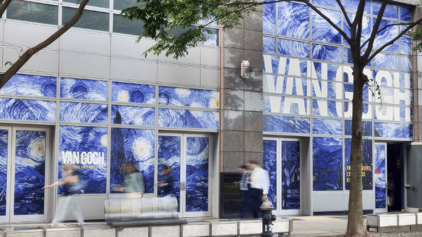 A building exterior featuring large window graphics of Vincent van Gogh’s artwork, with pedestrians walking past on the sidewalk.