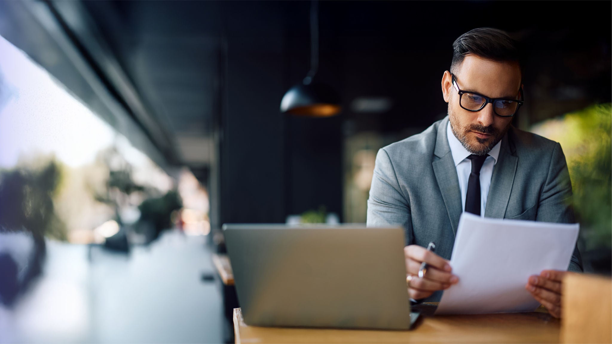 A professional man in a suit reviews printed documents beside a laptop in a modern workspace, focused on analyzing information.
