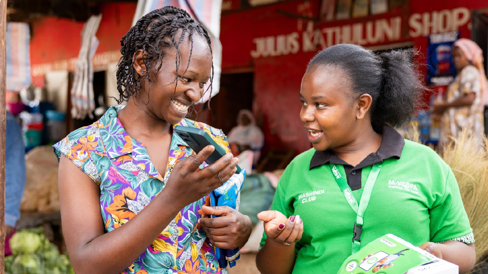 A customer and an M-KOPA representative reviewing product information on a smartphone at an outdoor market.
