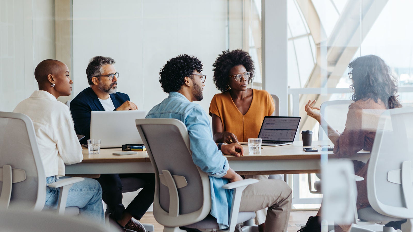 A group of professionals seated around a table in a modern office, discussing and collaborating with laptops and documents.