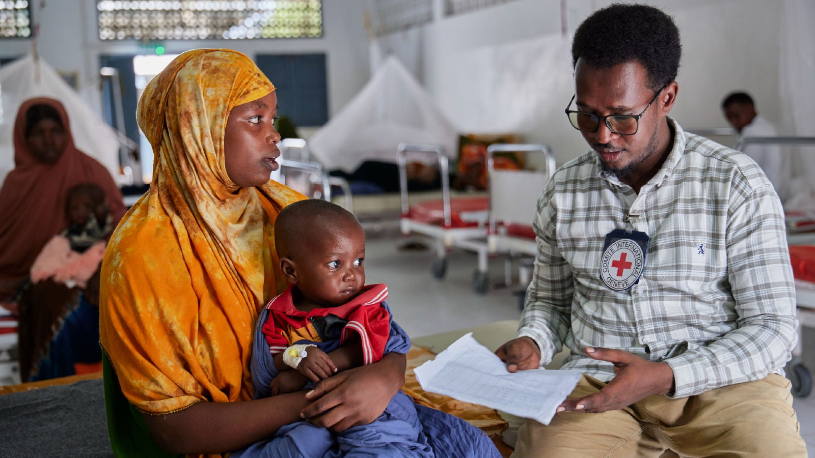 A woman in an orange headscarf holding a young child sits with an ICRC staff member in a medical facility, reviewing documents.