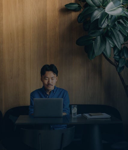 Professional working on a laptop at a café table beneath a large indoor plant.