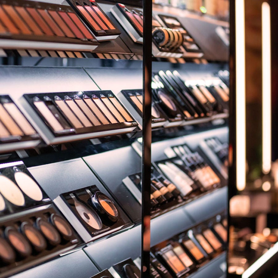 Close-up of makeup products displayed on sleek black store shelves, including eyeshadow palettes, powders, and foundation compacts, illuminated by warm lighting.