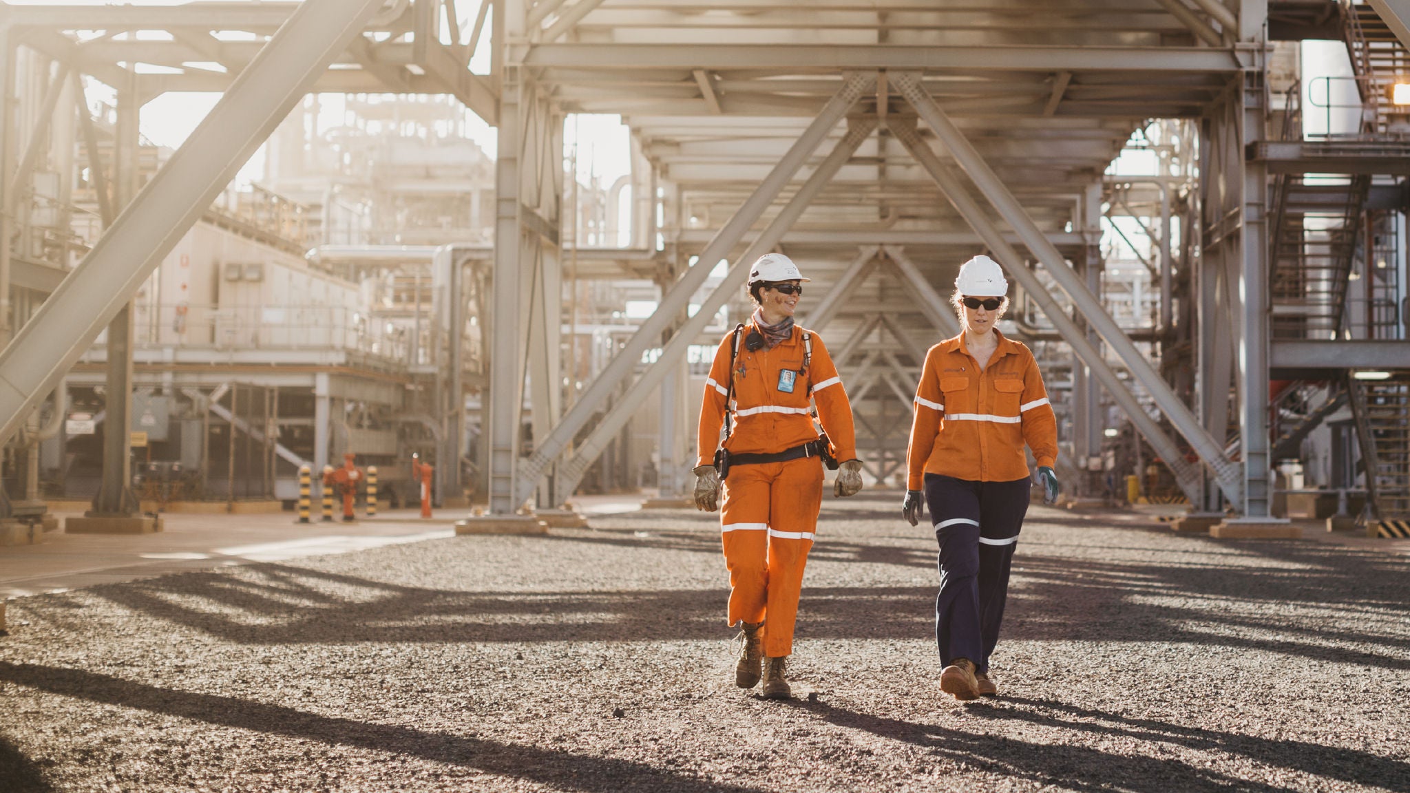 Two workers in orange protective uniforms walk across a gravel-covered area beneath large metal industrial structures.
