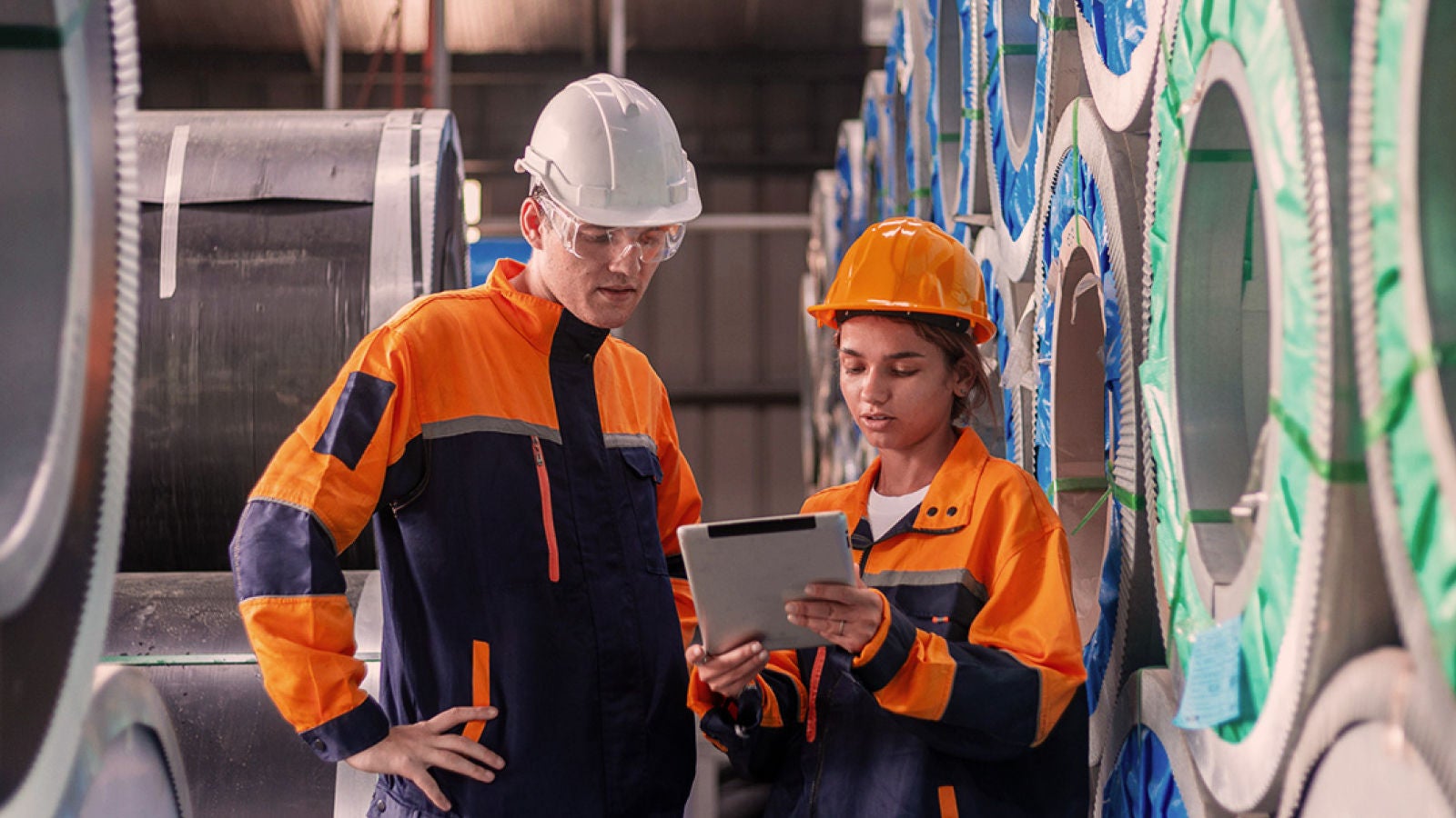 Two industrial workers in safety helmets and orange protective uniforms reviewing information on a tablet beside stacked metal coils in a manufacturing facility.