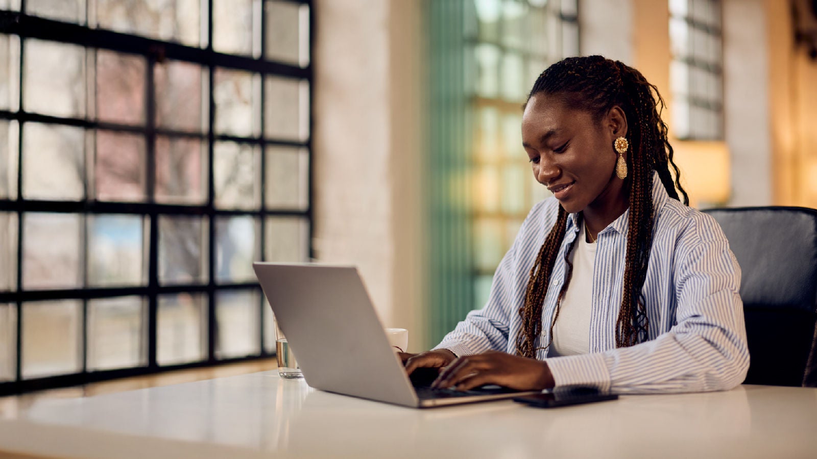 A woman seated at a desk typing on a laptop beside a cup and smartphone, in a bright office with large windows, representing focus and data analysis.