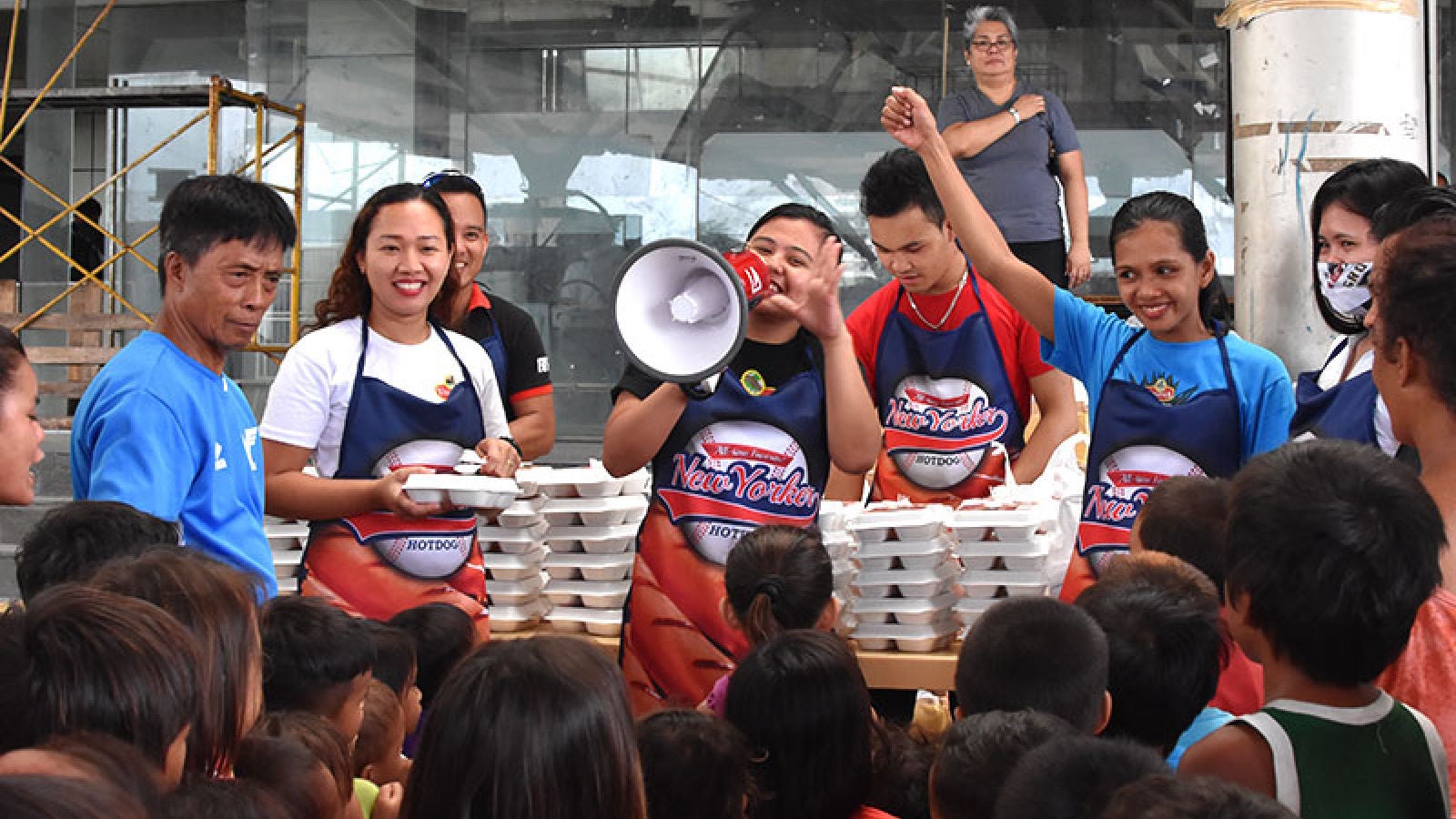 Community volunteers wearing Virginia Food aprons distribute boxed meals to a crowd while speaking through a megaphone.