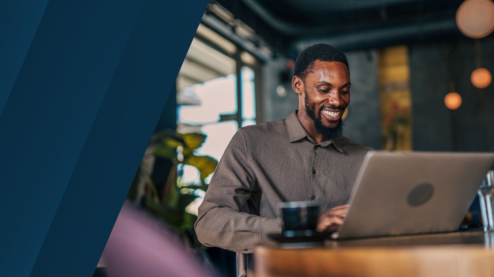 Smiling man working on a laptop at a café-style table with plants and warm lighting in the background.
