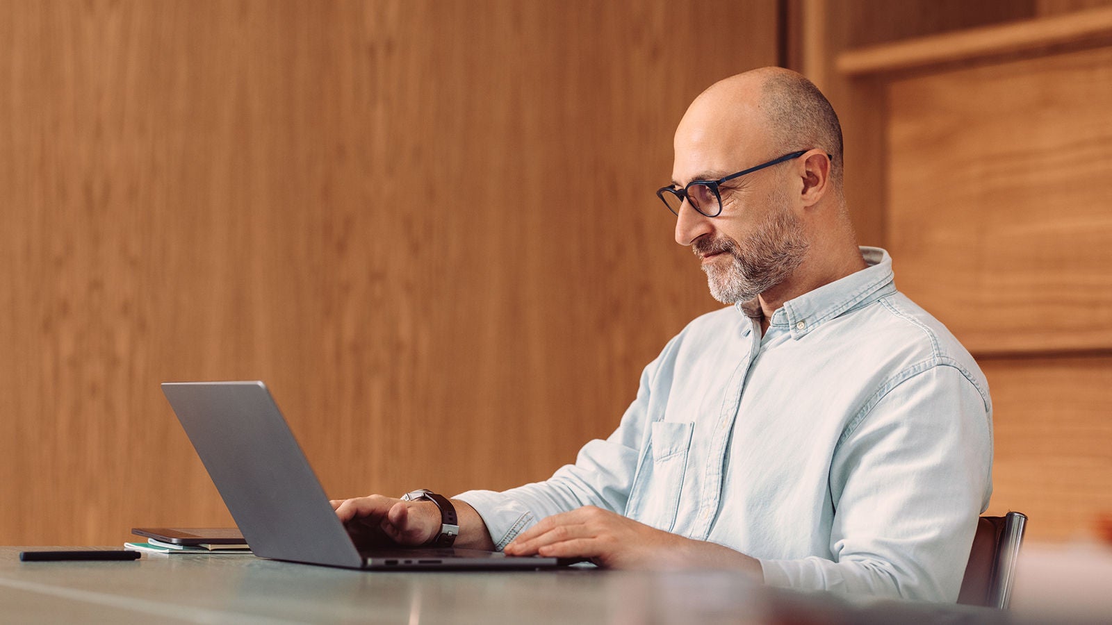 A man with glasses and a beard works on a laptop at a desk in front of a wooden wall panel.