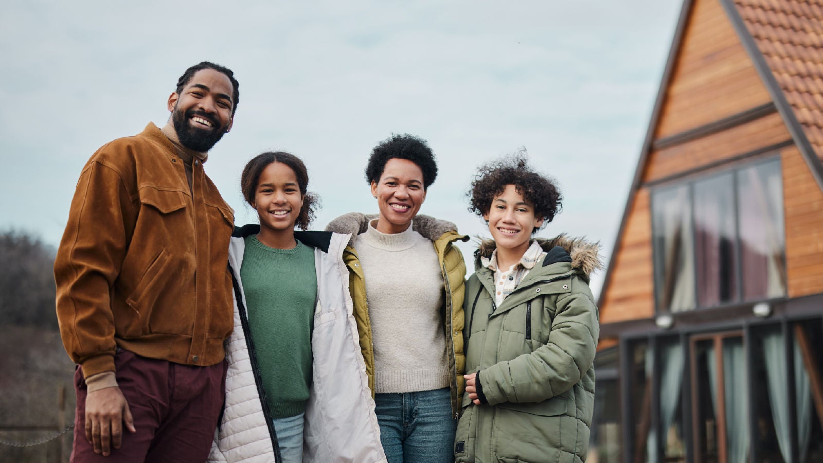 A family of four standing outside a wooden A-frame house, smiling together in cold-weather clothing.