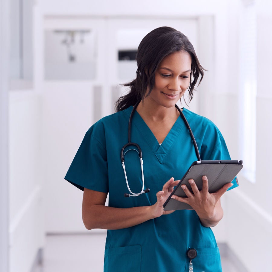 A woman in a scrub suit is holding a tablet computer, focused on her work in a healthcare setting.
