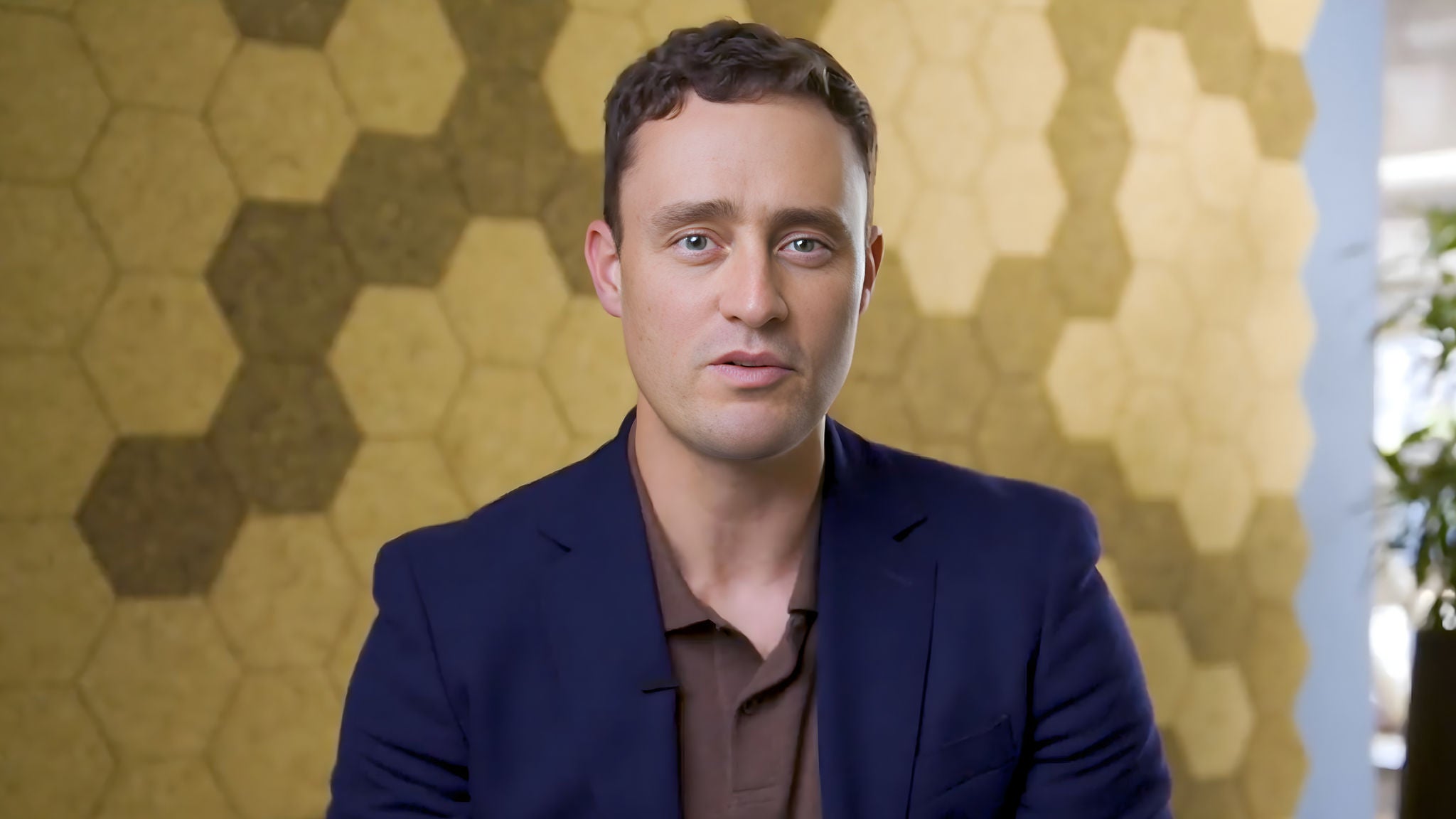 Man in business attire speaking in front of a wall with a hexagonal pattern background.