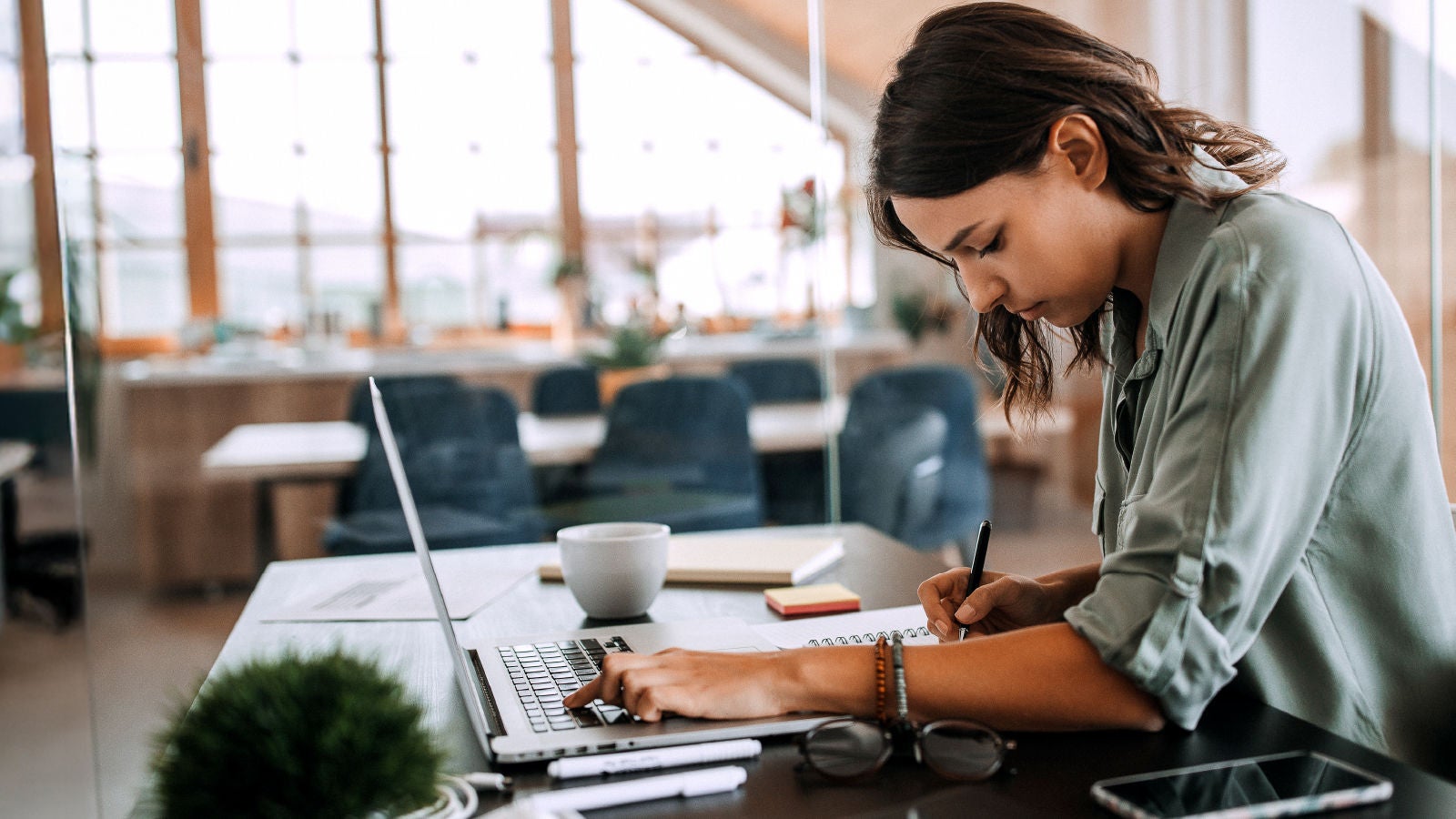A woman in a light green shirt working at a laptop, writing in a notebook with a coffee cup, smartphone, and glasses nearby in a modern, sunlit office space.