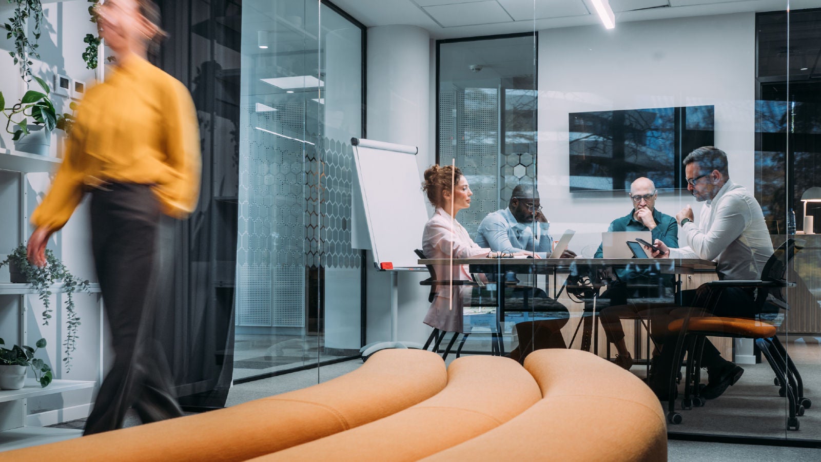 Four professionals sit together in a glass-walled meeting room, focused on laptops and printed materials, while a person in a yellow blouse walks past the room in the foreground.