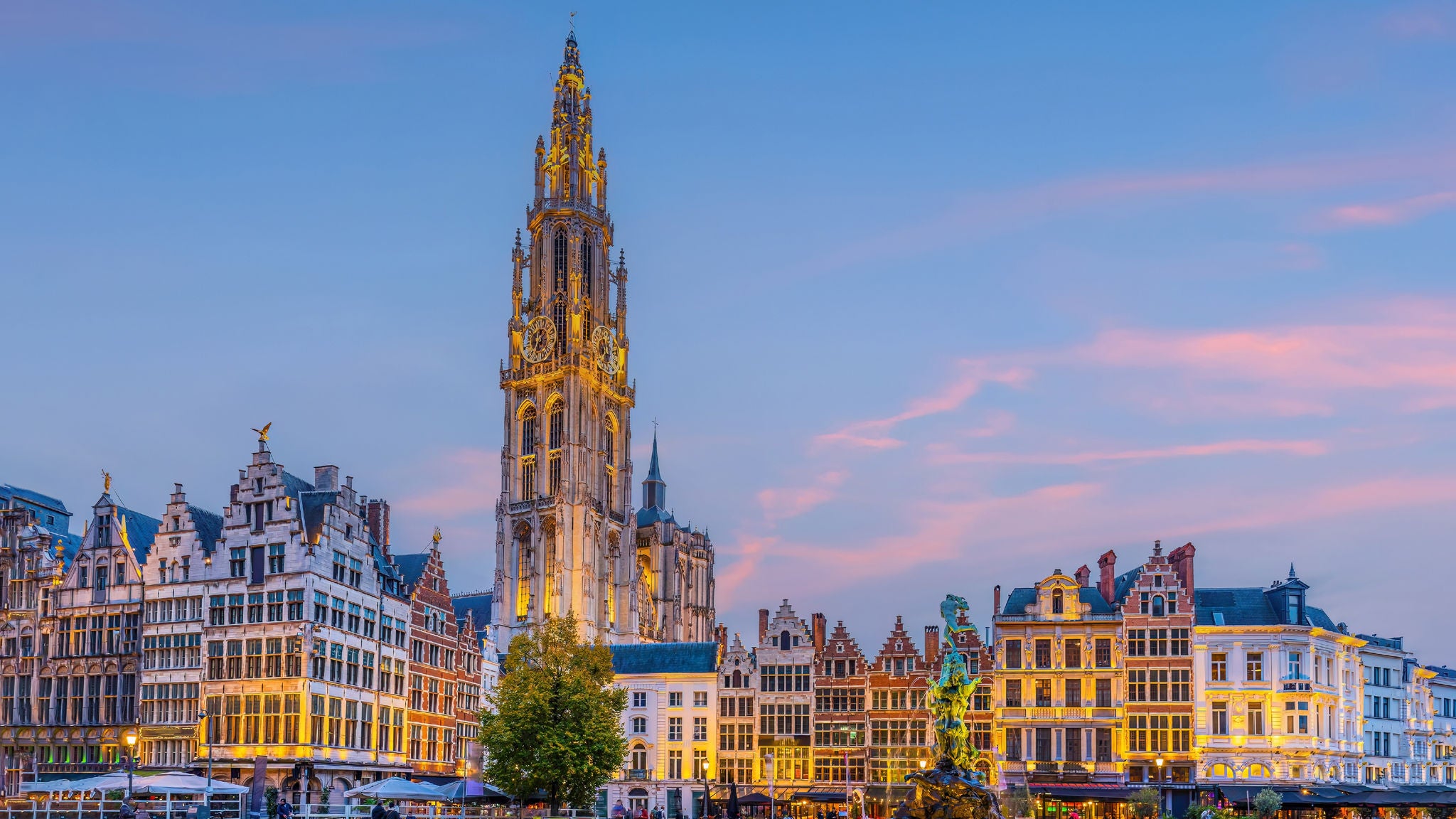 Historic city center of Antwerp, Belgium, with illuminated guild houses and the Cathedral of Our Lady at dusk under a blue and pink sky.