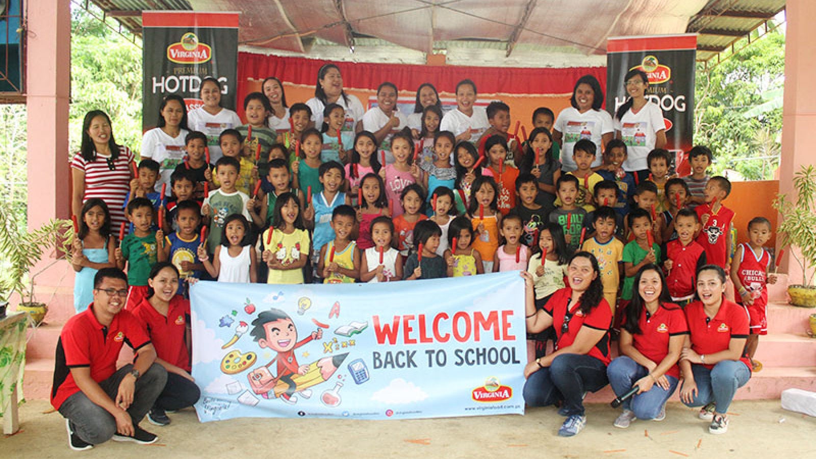 Large group of children and volunteers posing together while holding a “Welcome Back to School” banner during a community outreach event.