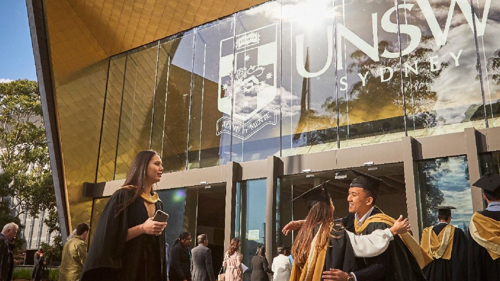 Graduates in caps and gowns greeting each other outside a UNSW Sydney building with large glass windows.