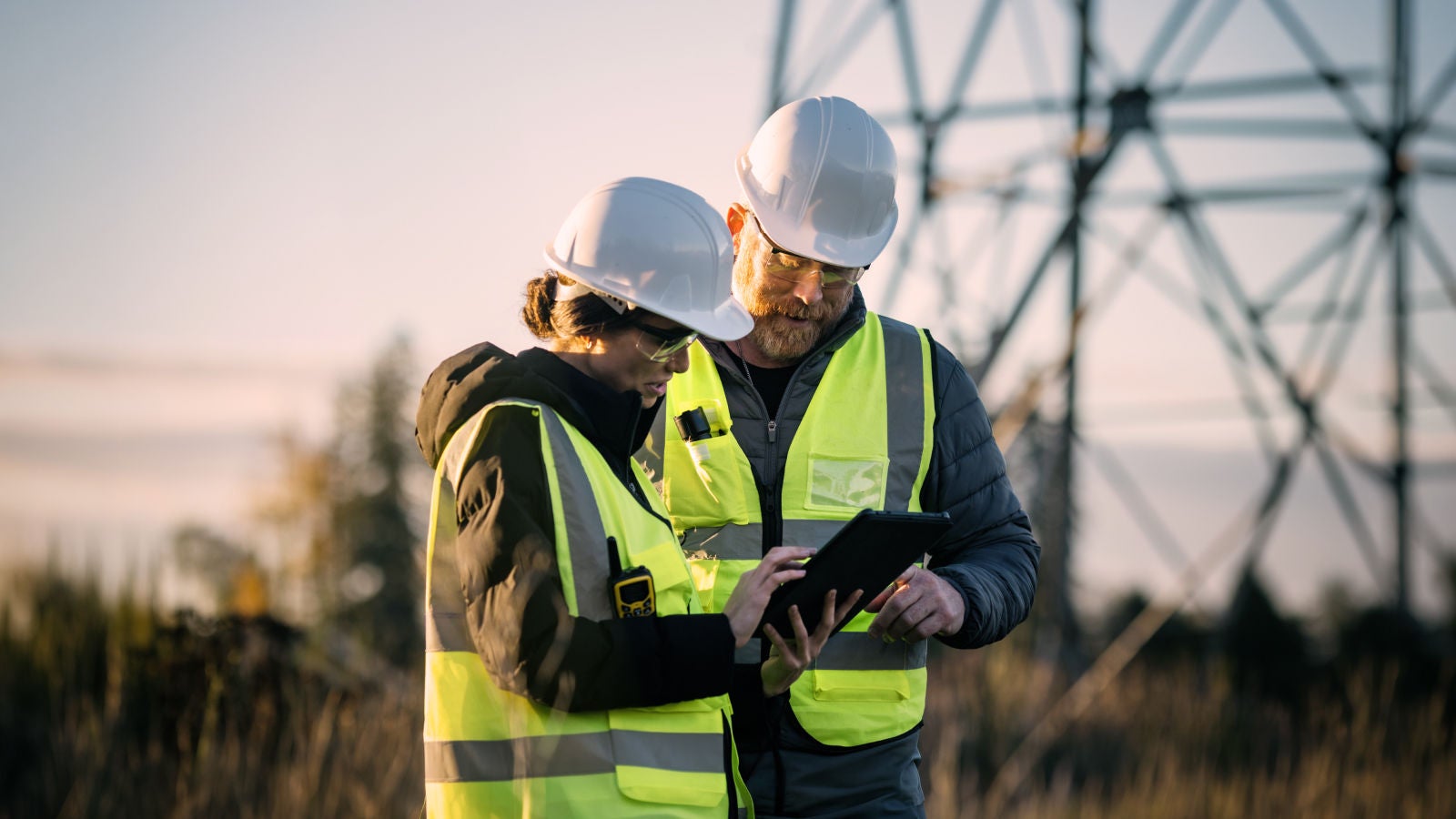 Two utility workers in safety vests and hard hats reviewing a tablet near power transmission towers at sunset.