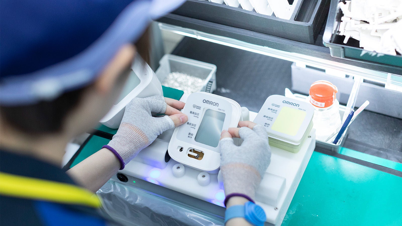 A worker wearing gloves carefully assembling or inspecting an Omron blood pressure monitor device on a production line.