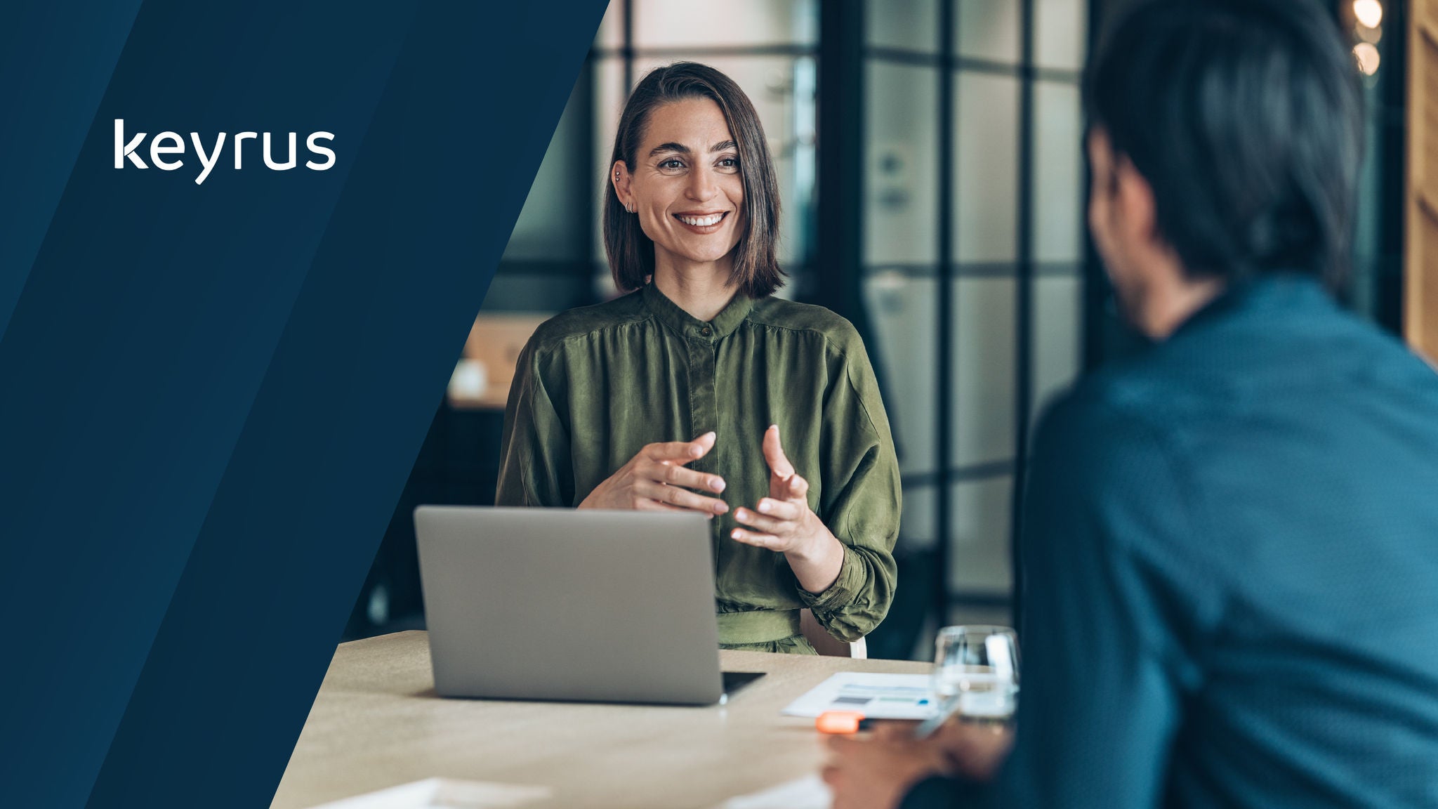 Keyrus Logo displayed on the left. A professional woman smiling and gesturing while speaking at a desk with a laptop, facing another person in a modern office setting.
