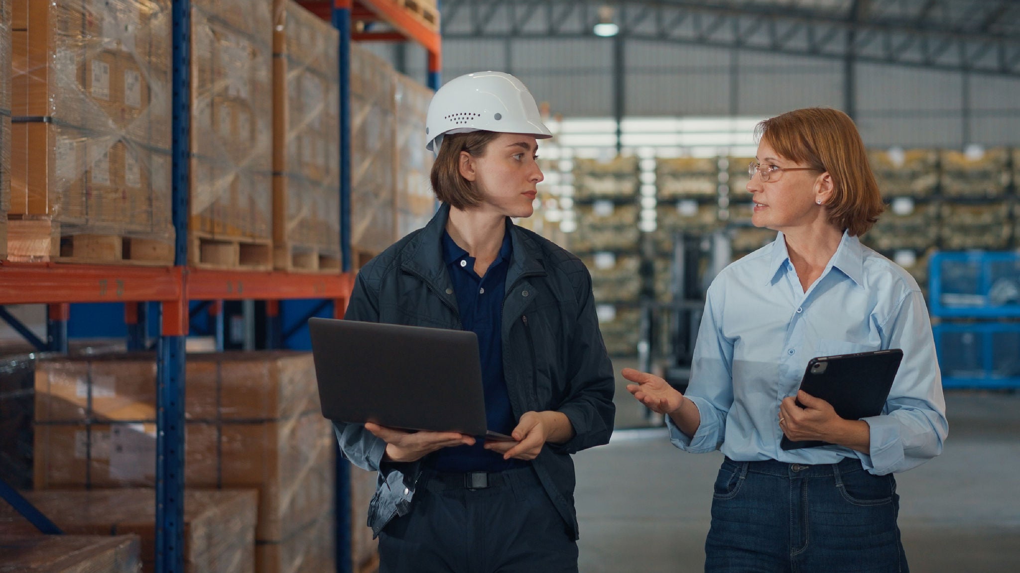 Two professionals wearing safety gear walking through a warehouse while reviewing inventory using a laptop and a tablet, with stacked pallets visible in the background.