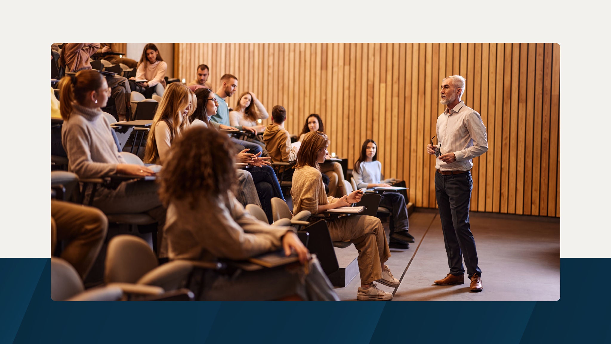 A professor stands at the front of a lecture hall, engaging with students seated in tiered rows. The setting features a wooden wall backdrop and a professional academic environment.
