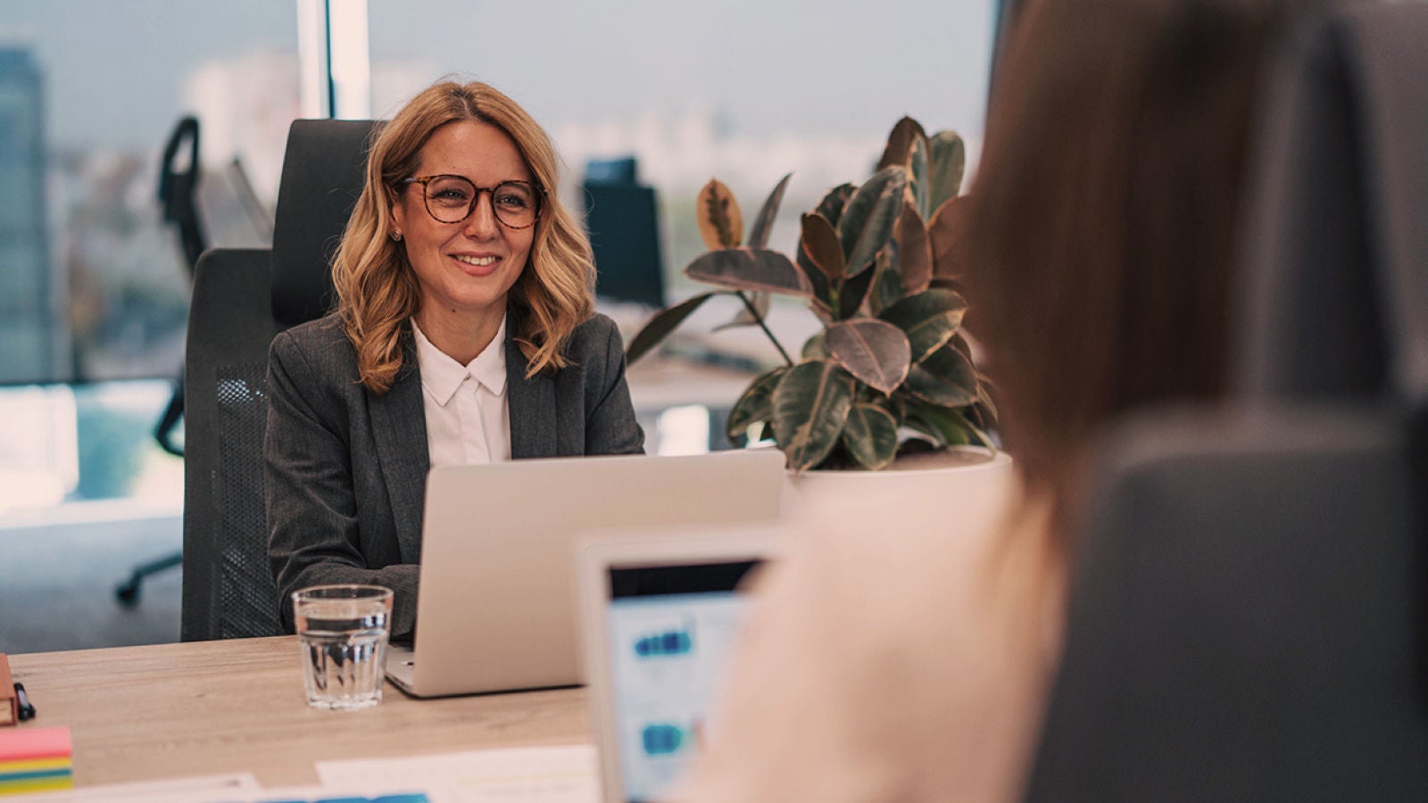 Businesswoman in an office meeting using a laptop while speaking with a colleague across the desk.